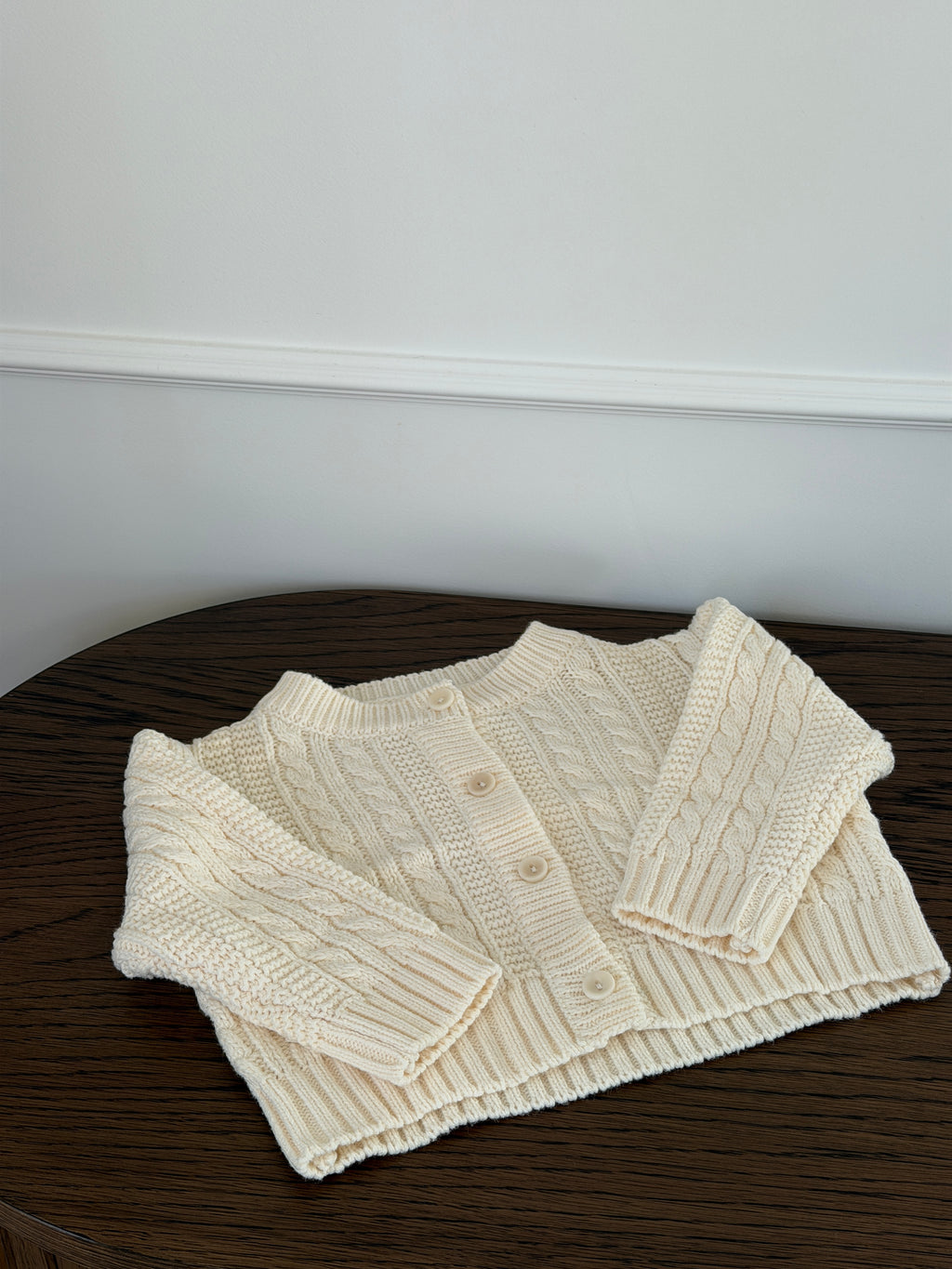 Creme kabelstrikket cardigan til baby liggende på træbord