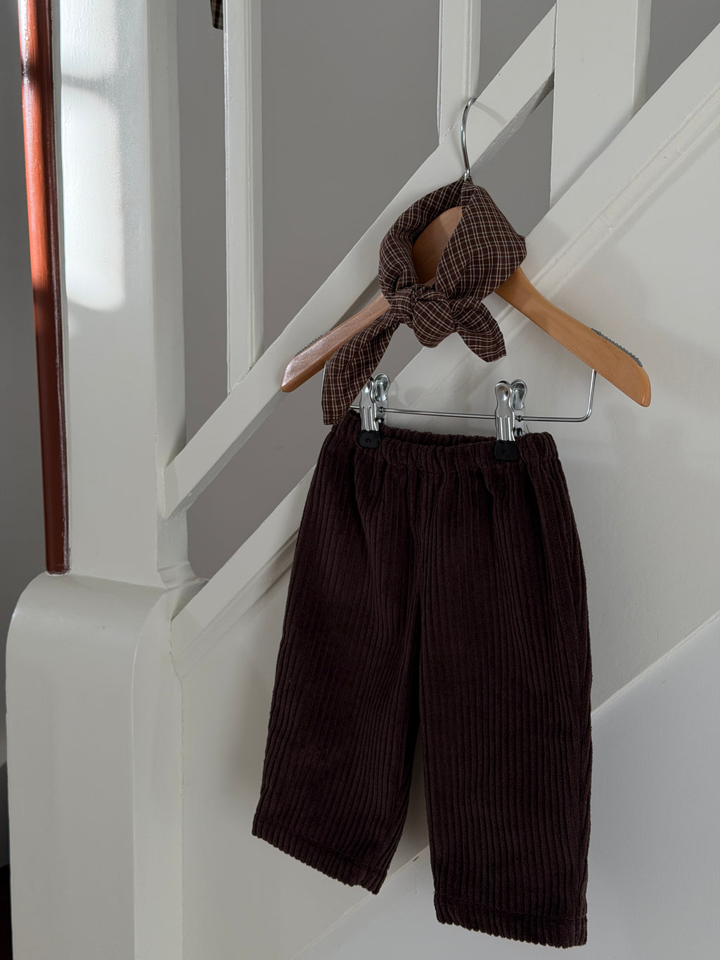 Brown velvet trousers and a brown checked scarf on a hanger by a white staircase