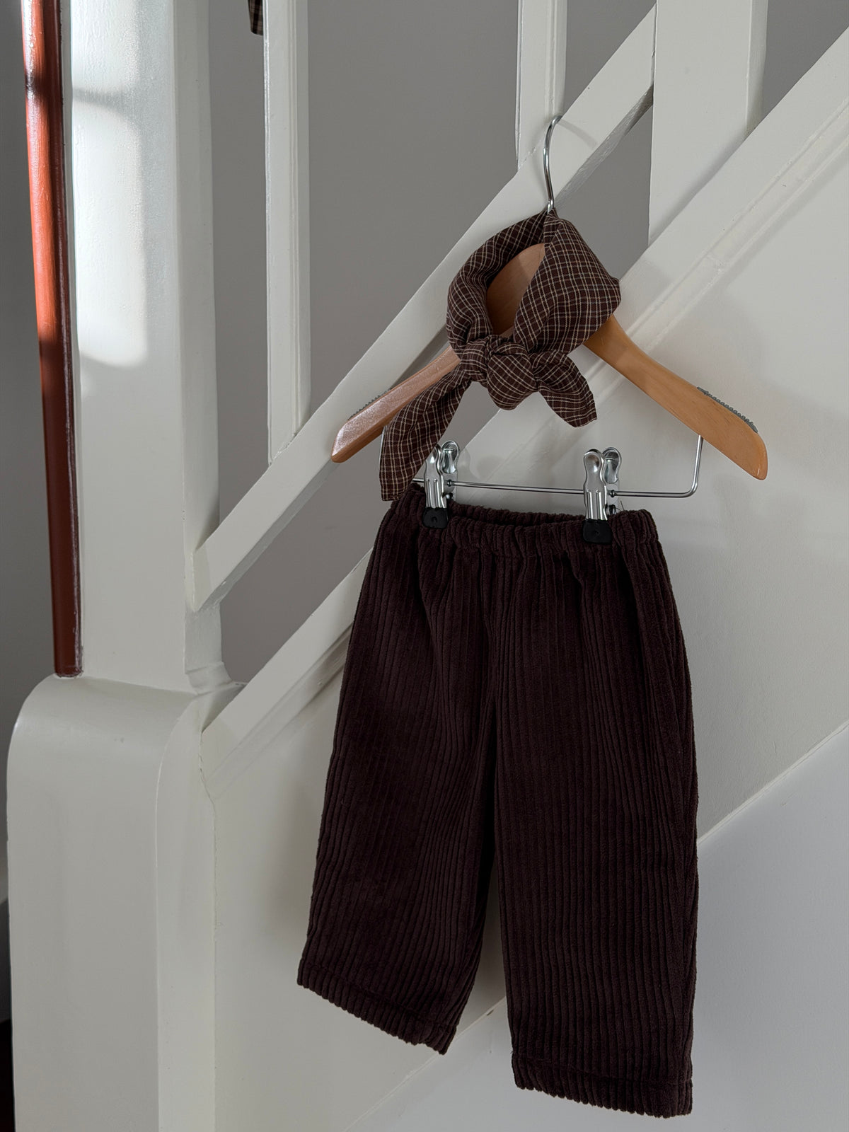 Brown velvet trousers and a brown checked scarf on a hanger by a white staircase