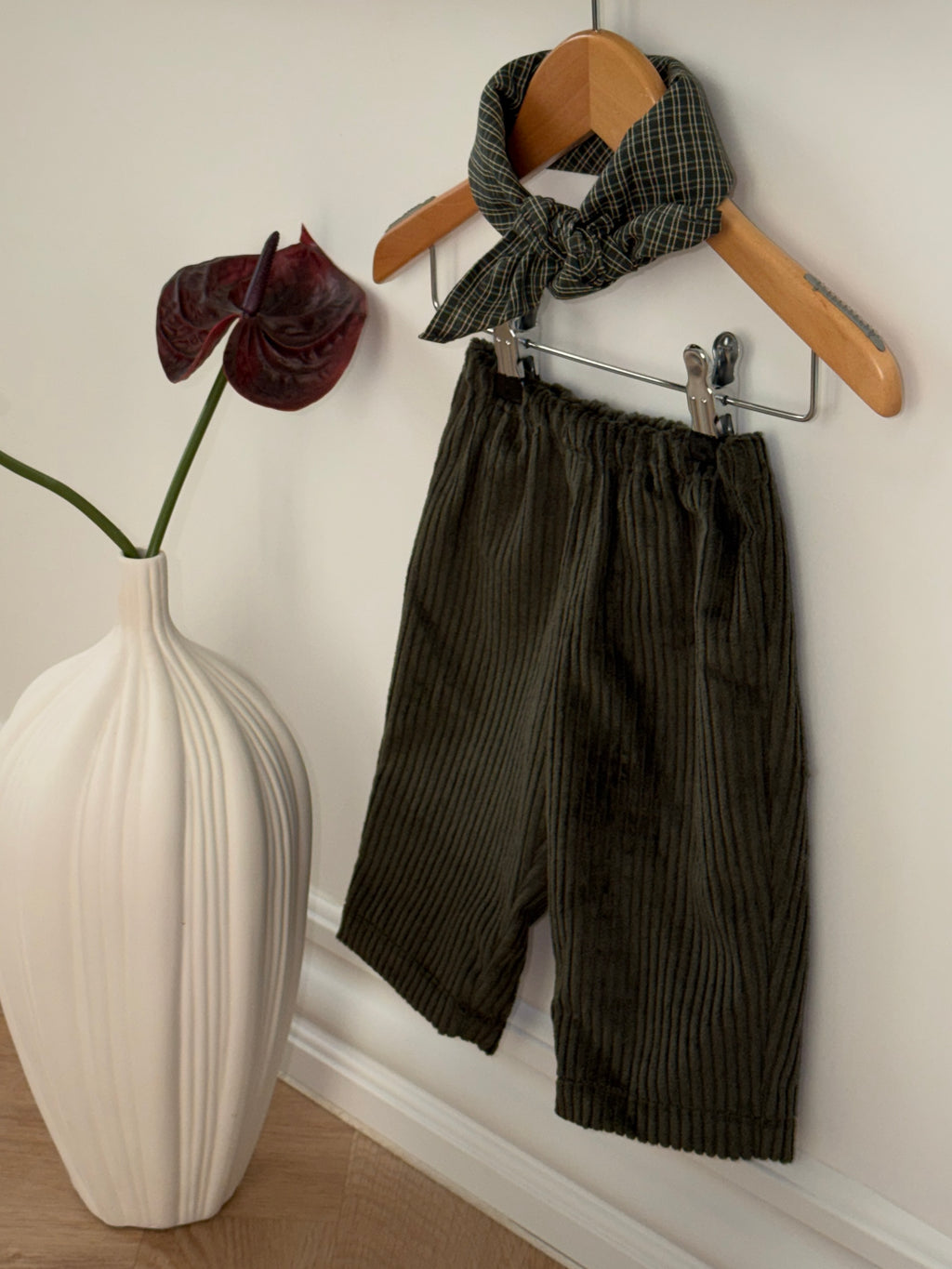 Green velvet trousers and a green checked scarf on a hanger by a white vase