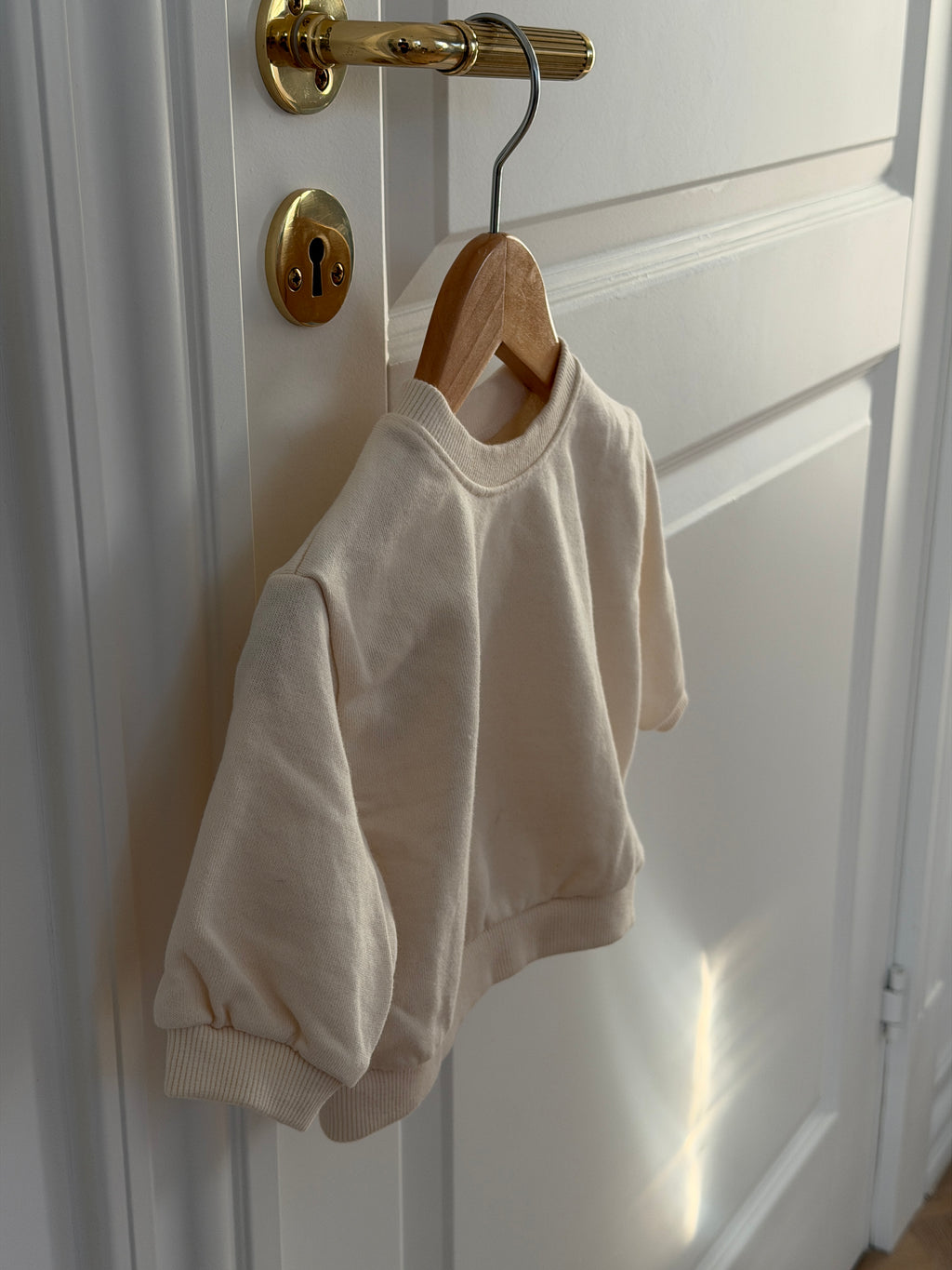 Cream crew-neck sweatshirt for baby on a hanger on a door