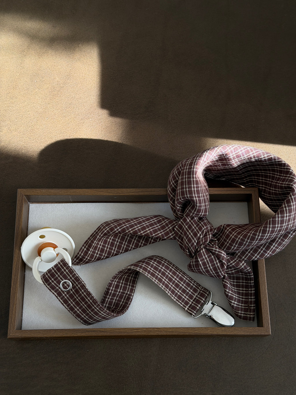 Pecan scarf and dummy clip in reddish-brown check with a dummy in a wooden tray in sunlight