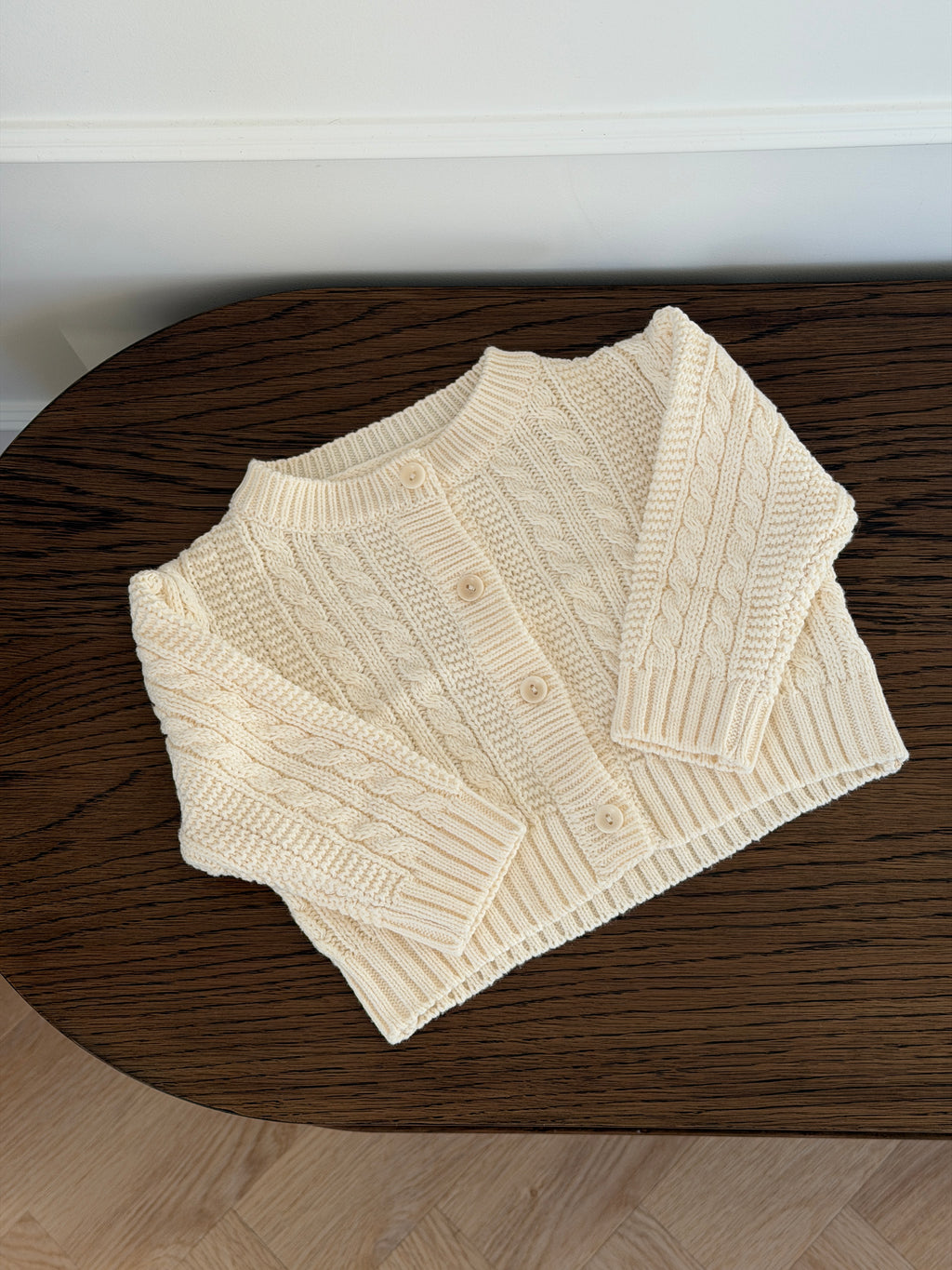 Creme kabelstrikket cardigan liggende på træbord