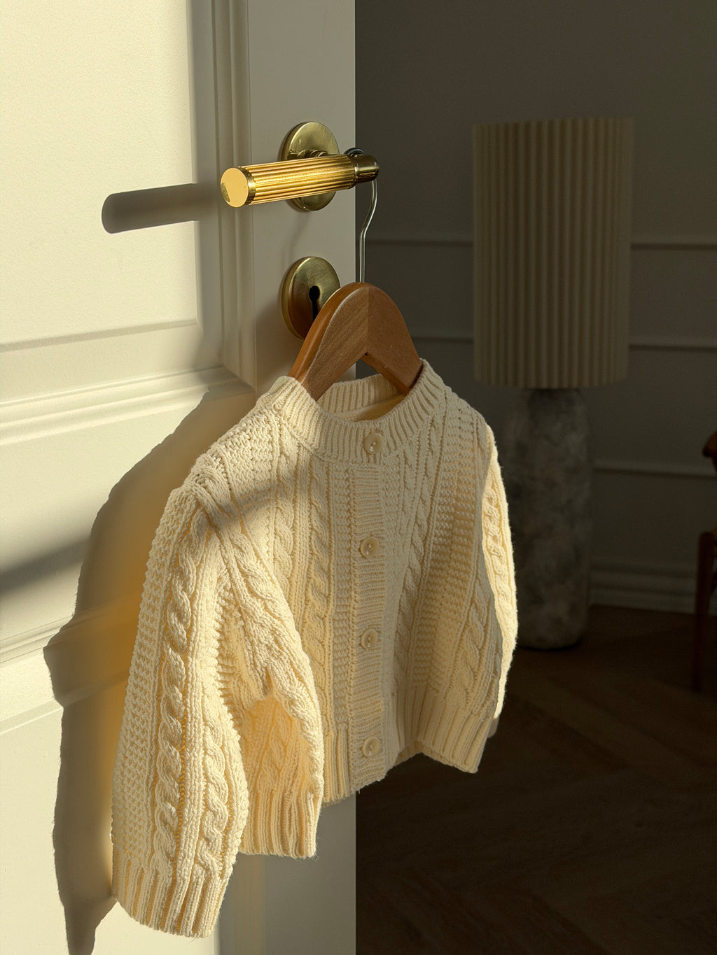 Creme kabelstrikket cardigan til baby på bøjle på dør